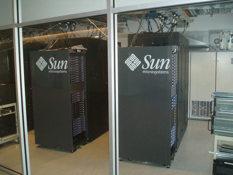 Two Sun Microsystems server racks stand behind glass walls in a data center, their blade servers stacked floor to ceiling, cables running overhead — the hardware that powered the early internet.